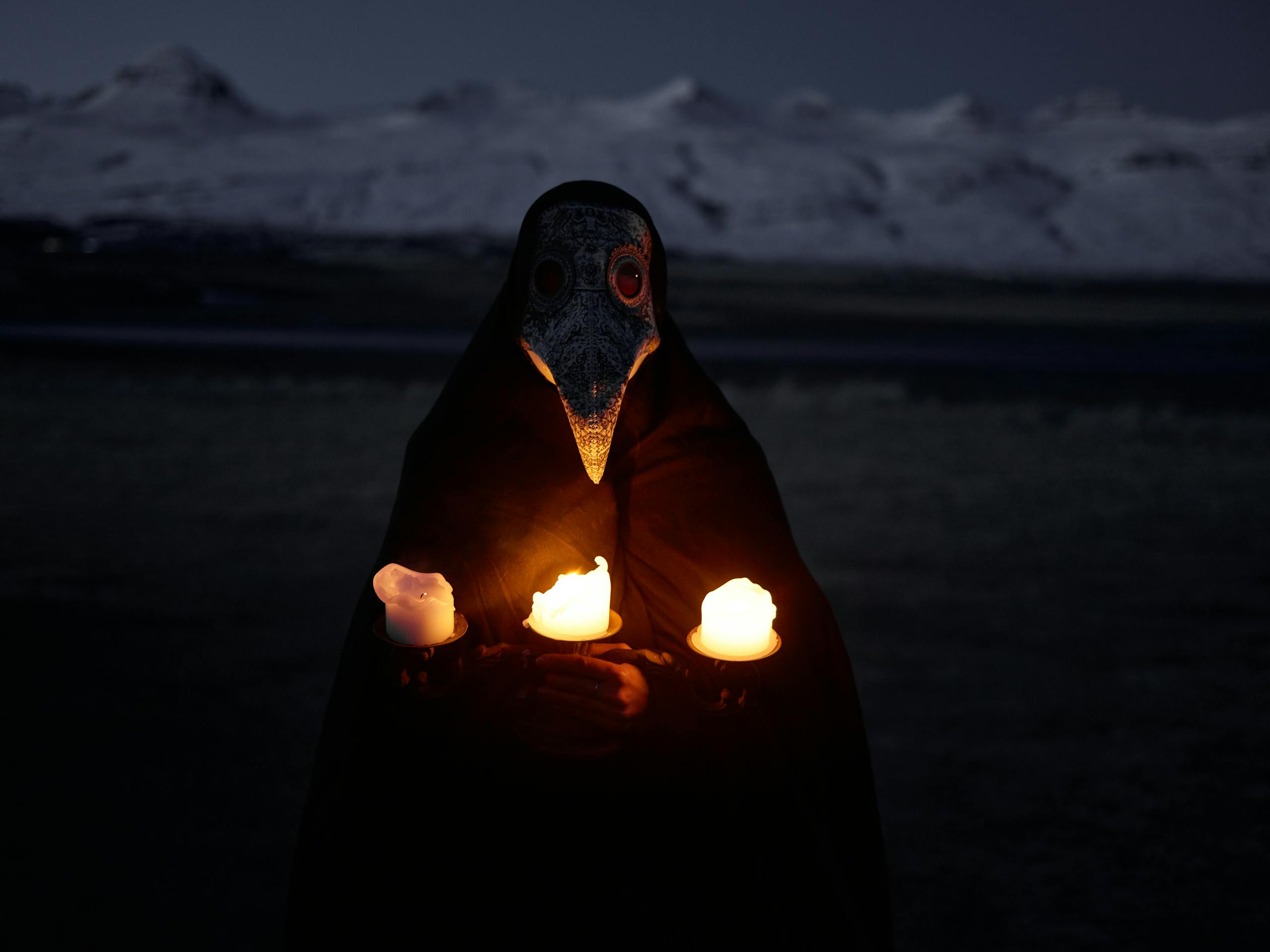 Unrecognizable person in plague mask standing in front of snowy hills at night and holding burning candles while looking at camera